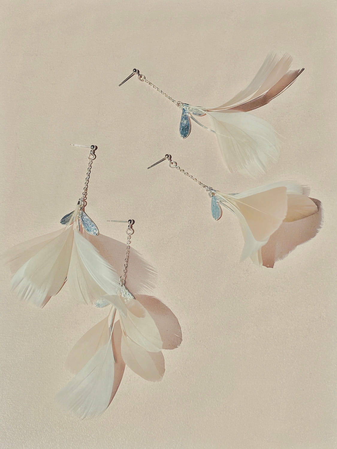 The Marie Earrings from Noémiah feature lightweight, high-quality feathers adorned with small blue gemstones. Set against a neutral, textured background, the feathers gracefully cascade with metal chain attachments and are elegantly finished with gold-filled hooks.