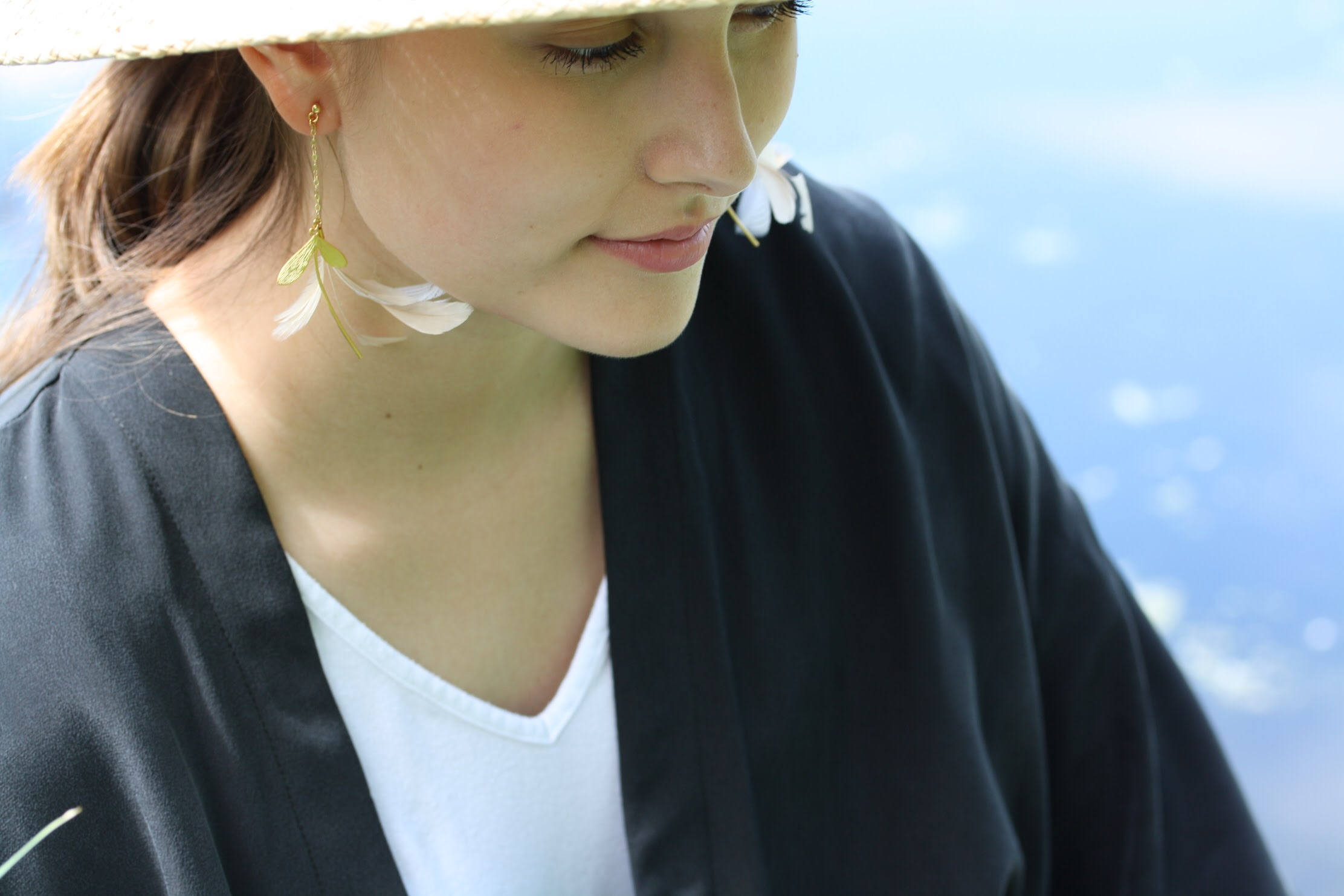 A woman wearing the Marie Earrings by Noémiah, which feature lightweight floral designs on gold-filled hooks, looks downward while dressed in a black and white outfit. The serene blurred blue background suggests water or sky.