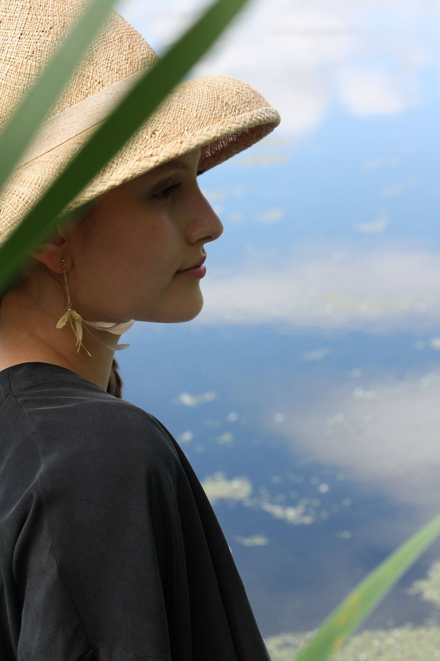 A person wearing a large straw hat and a black shirt is pictured in profile next to a body of water. Tall green grass partially frames the image, and the person adorns themselves with the elegant Marie Earrings by Noémiah, featuring lightweight, gold-filled hooks that dangle gracefully in the shape of leaves.