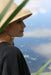 A person wearing a large straw hat and a black shirt is pictured in profile next to a body of water. Tall green grass partially frames the image, and the person adorns themselves with the elegant Marie Earrings by Noémiah, featuring lightweight, gold-filled hooks that dangle gracefully in the shape of leaves.