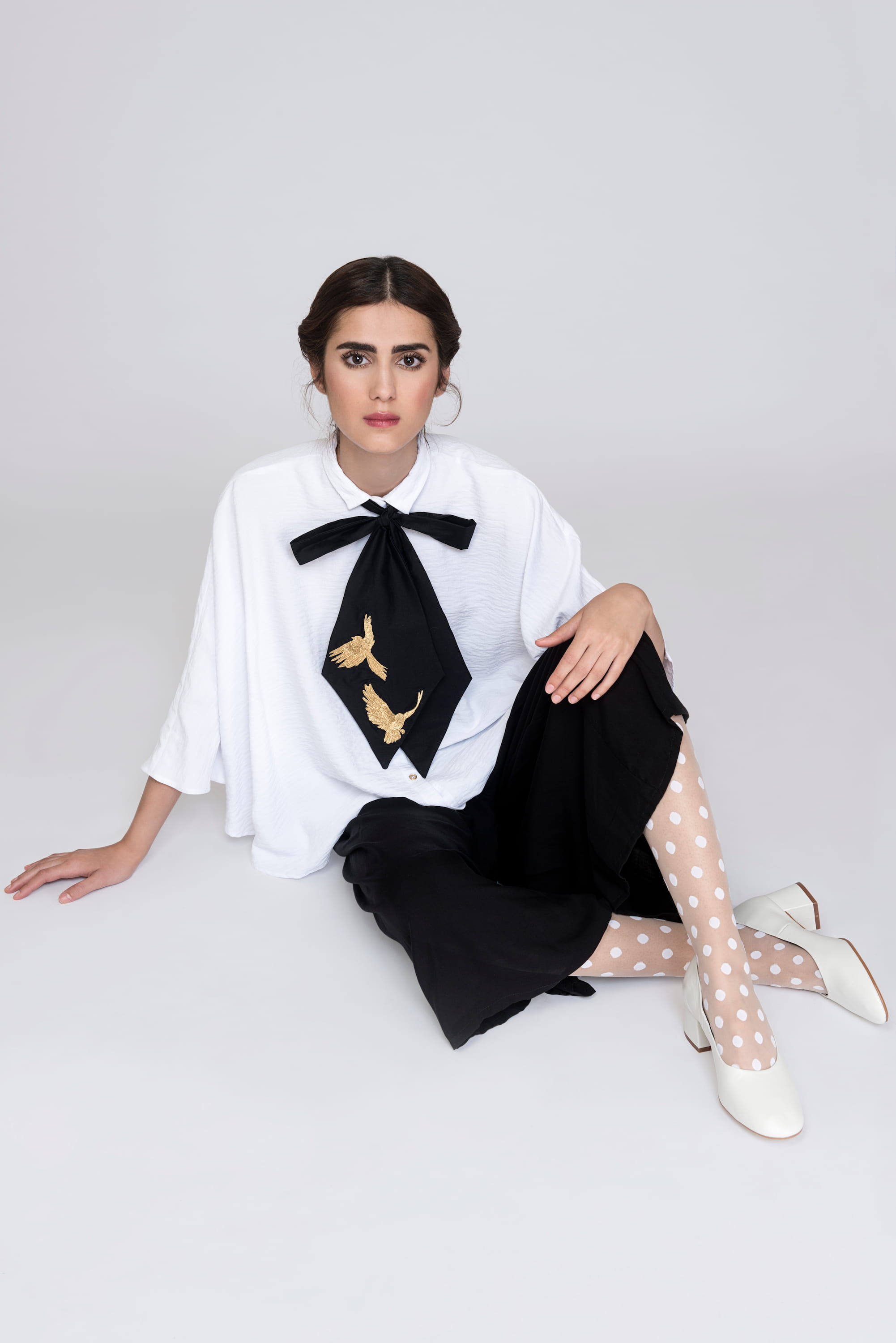 A woman sits elegantly on the ground against a simple backdrop, wearing the Marie Shirt by Noémiah. The white blouse features gold metal buttons and is complemented by a black bow tie embellished with golden birds. She pairs it with a black skirt, polka dot stockings, and white heels. Her hair is tied back, enhancing her sophisticated appearance.