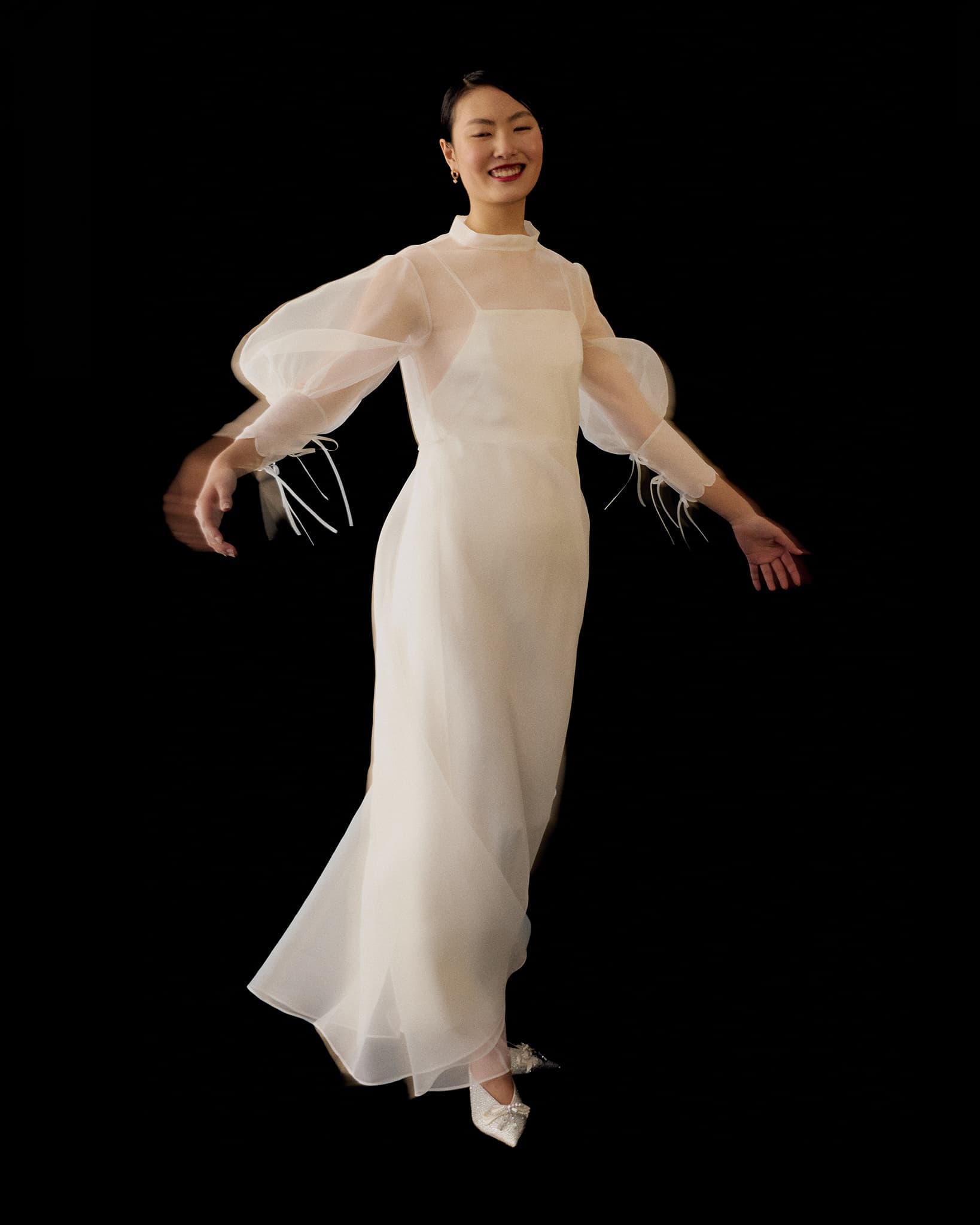 A woman smiles and twirls in the Marine Dress by Noémiah, a long sheer silk organza gown with puffy lantern sleeves. She pairs it with white pointed shoes and simple earrings against a black background.