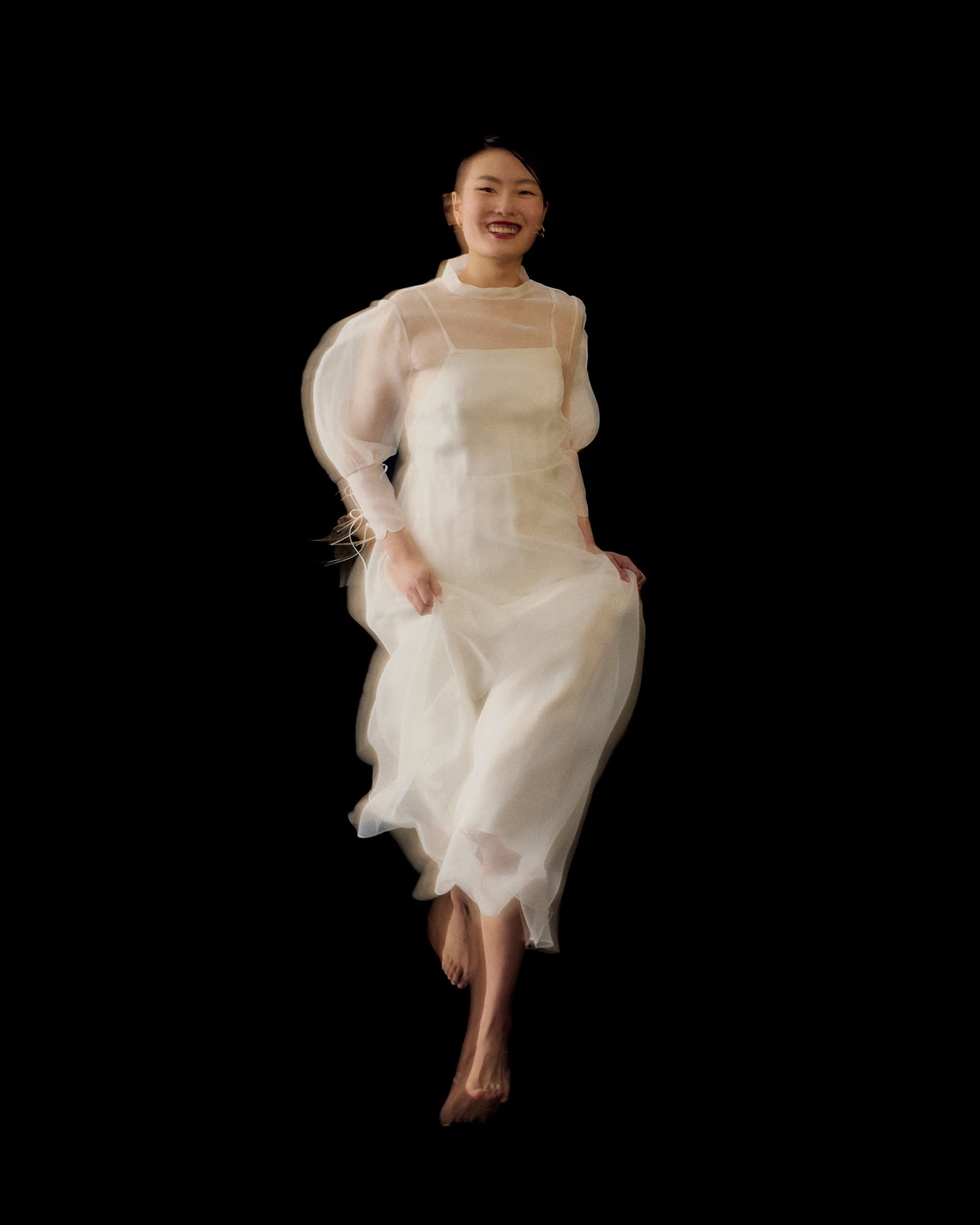 A woman wearing the Noémiah Marine Dress, a sheer white silk organza design, smiles mid-jump against a black background, her bare feet lifted slightly off the ground.