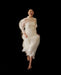 A woman wearing the Noémiah Marine Dress, a sheer white silk organza design, smiles mid-jump against a black background, her bare feet lifted slightly off the ground.