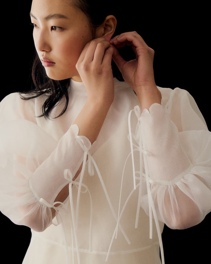A person in the Noémiah Marine Dress, a sheer white blouse with long lantern sleeves and ribbon accents, adjusts an earring while gazing to the side against a black background.
