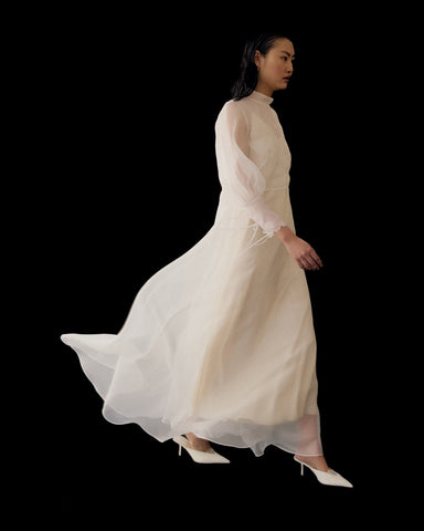 A woman in the Noémiah Marine Dress—long, flowing silk organza with lantern sleeves—gracefully walks in white heels against a black background, the elegant dress billowing behind her.