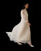 A woman in the Noémiah Marine Dress—long, flowing silk organza with lantern sleeves—gracefully walks in white heels against a black background, the elegant dress billowing behind her.