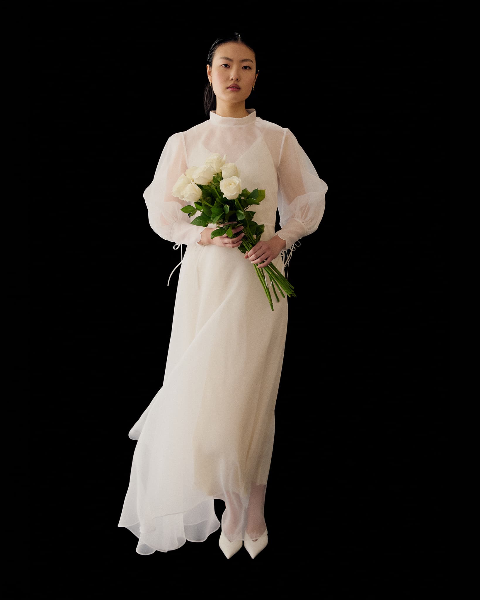 A woman wears the Marine Dress by Noémiah, a long sheer white silk organza piece with lantern sleeves. She holds white roses with greenery, stands before a plain black background in white shoes, and has a calm expression.