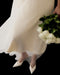 Wearing the Marine Dress by Noémiah—a flowing silk organza dress with lantern sleeves—a person holds white roses and wears white pointed lace-detailed shoes, standing against a black background.