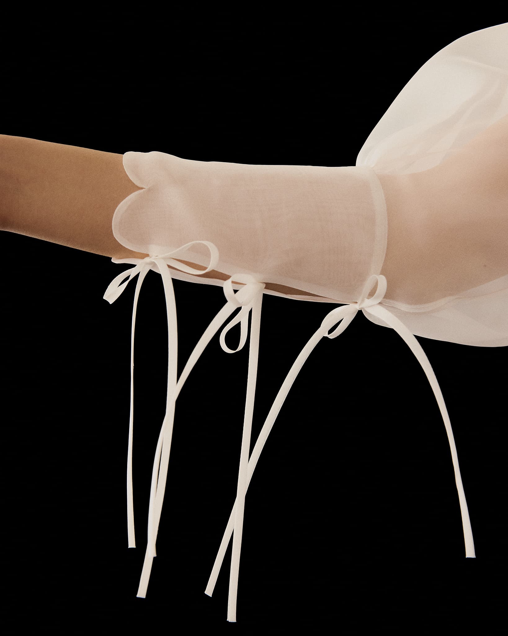 A sheer, white glove with long ribbon ties and delicate bows at the wrist and forearm is worn against a black background, echoing the elegance of the Noémiah Marine Dress.