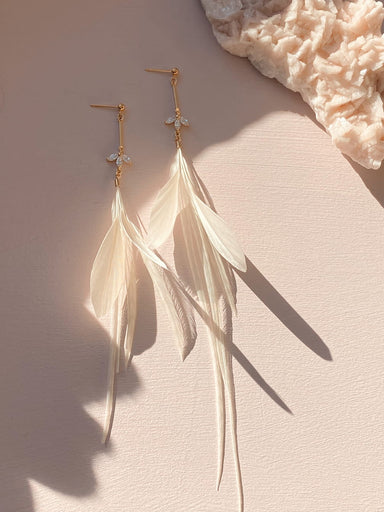 The Marlie Earrings by Noémiah feature gold accents and delicate white feathers enhanced by a subtle gold-plated bar, elegantly displayed on a light pink surface. Shadows add depth to the composition, with a textured beige element partially visible in the top right corner.