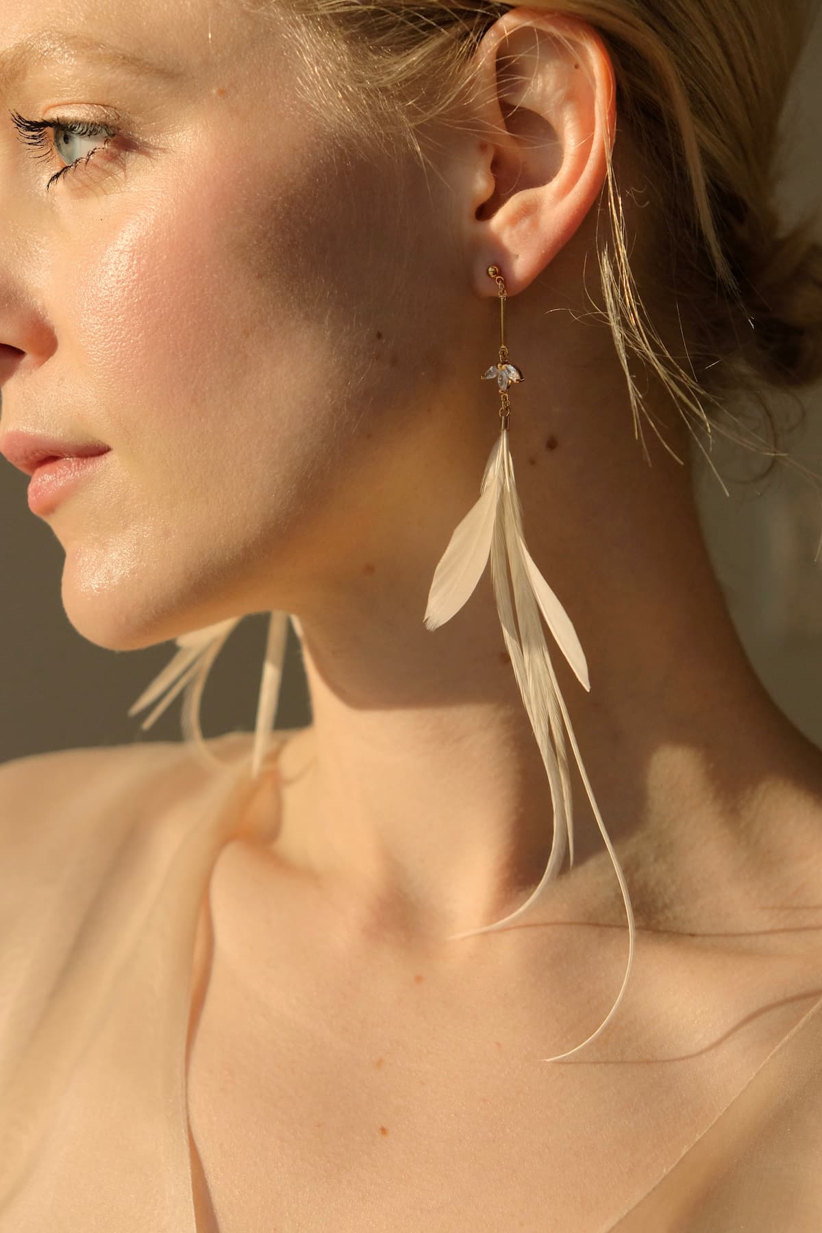 A woman is seen in profile wearing the Marlie Earrings by Noémiah, which feature white feathers. The sunlight casts a warm glow on her face and highlights the delicate, translucent fabric of her top, while a subtle cubic zirconia glimmers at her neckline.