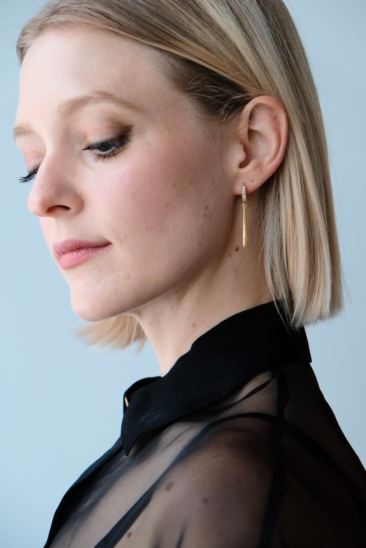 A person with short blonde hair and a side part wears a sheer black top and the Maude Earrings by Noémiah, standing against a light blue background, looking downwards.