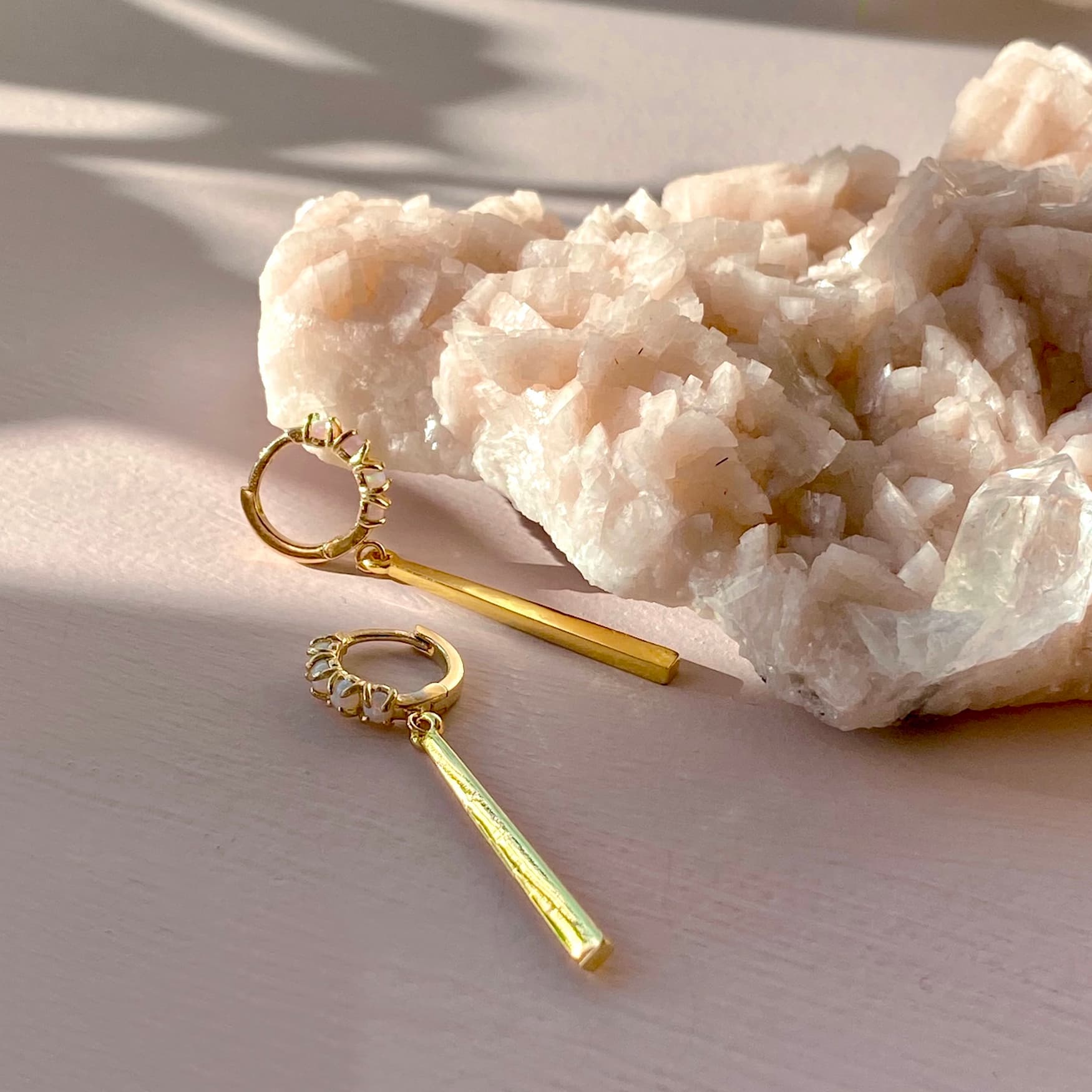 The Maude Earrings by Noémiah, featuring 14K gold-plated hoop earrings with long bar pendants, are elegantly showcased on a pale pink surface alongside a large, light pink crystal rock. The lustrous finish glistens under soft, natural light, creating delicate shadows.