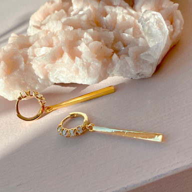 The Maude Earrings by Noémiah, elegantly designed with a 14K gold-plated hoop and bar motif, rest serenely on a surface next to a cluster of pale pink and white crystals. The soft lighting highlights both the earrings and the crystalline textures, creating a harmonious blend of luxury and natural beauty.