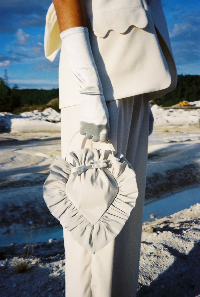 Wearing Noémiahs Frédérique Trousers — Bridal Edit, a person in an off-white suit and white gloves stands outdoors on rocky terrain under a blue sky, holding a heart-shaped ruffled bag. Only their torso and legs are shown.