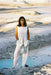 Wearing Noémiah’s Frédérique Trousers — Bridal Edit, a woman stands outdoors on white rocks by turquoise water, dressed in a sleeveless white suit with gloves and holding a silver pouch against a surreal landscape.