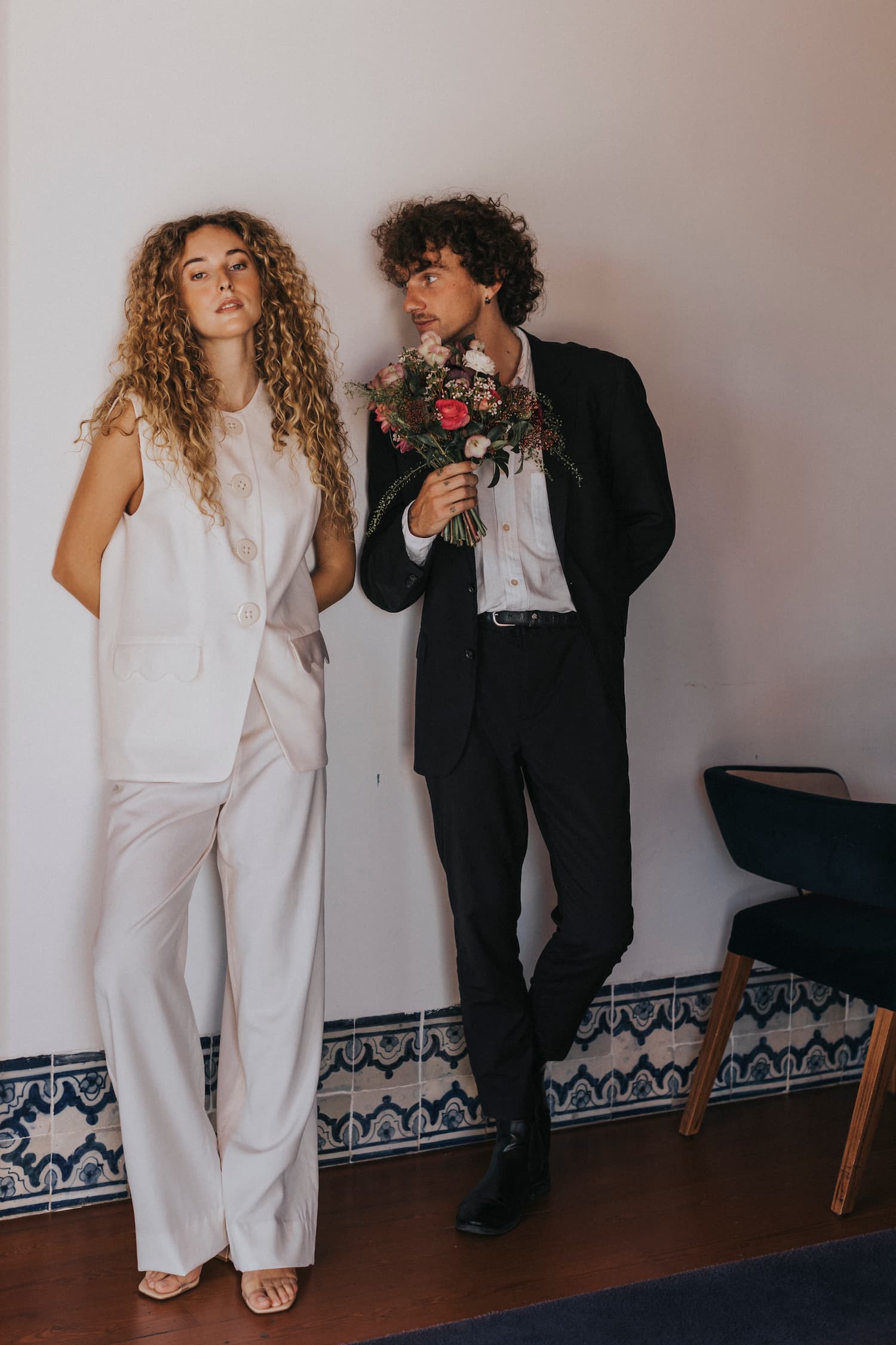 A woman with curly hair wearing the Maxime Vest by Noémiah, distinguished by its porcelain buttons, stands against a wall. Meanwhile, a man dressed in a dark suit leans toward her with a bouquet of flowers in hand. The room's base features decorative blue patterned tiles.