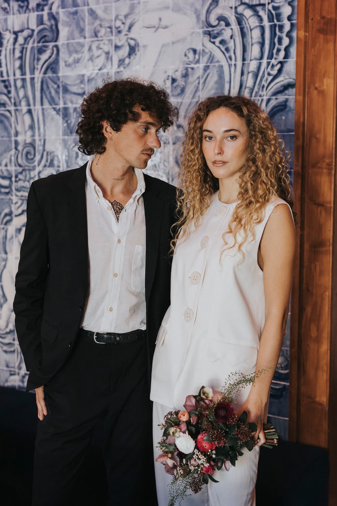 A man in a black suit and a woman wearing the Noémiah Maxime Vest, featuring elegant porcelain buttons, stand closely together. The woman holds a bouquet of flowers against a backdrop of a blue and white patterned wall.