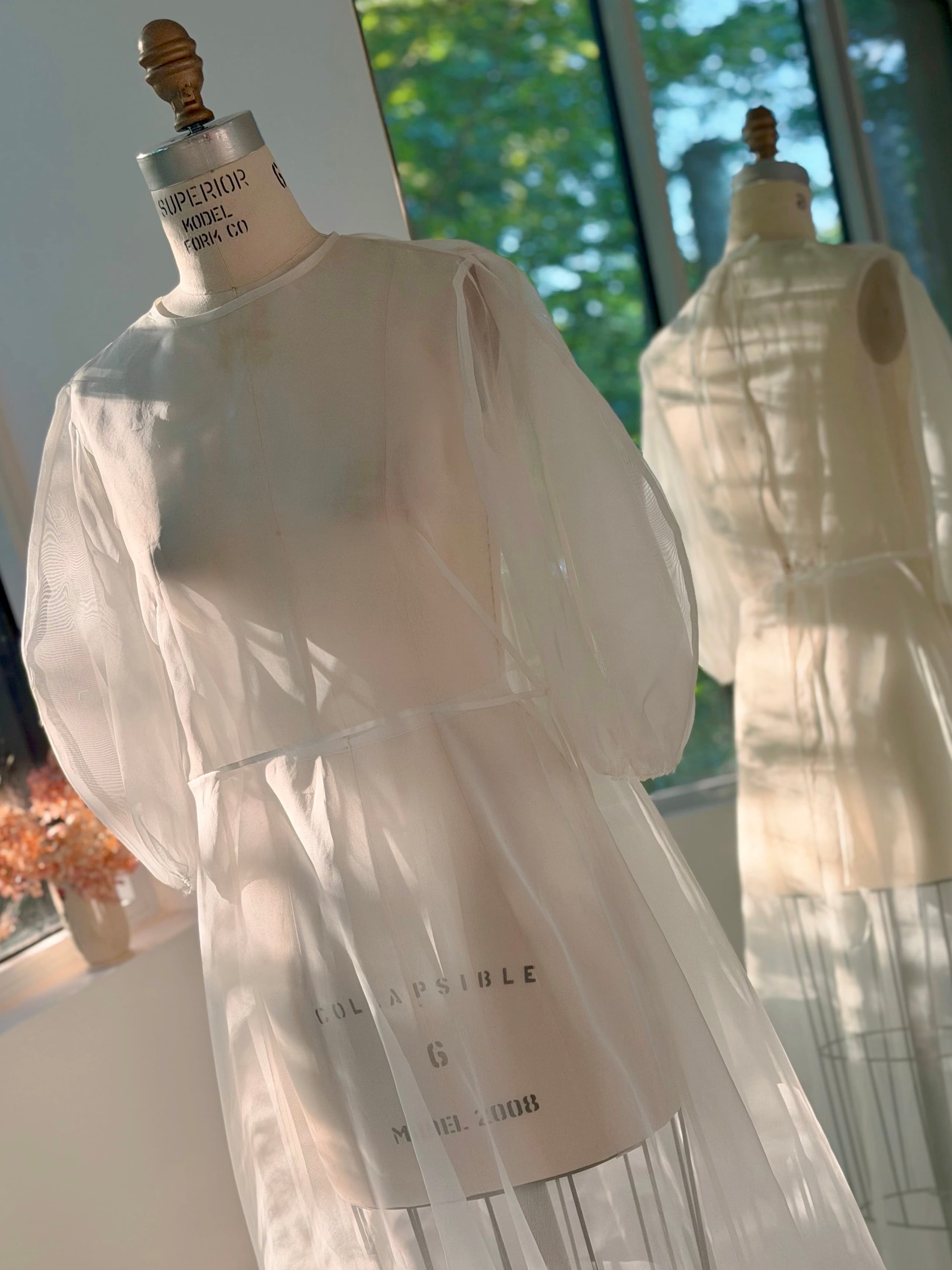 The Maya Dress by Noémiah, crafted from sheer white silk organza, is displayed on a dress form as sunlight pours through large windows, casting soft reflections on a nearby mirror. This vintage-inspired maxi dress features delicate puffed sleeves, bringing an ethereal touch to the elegant setting.