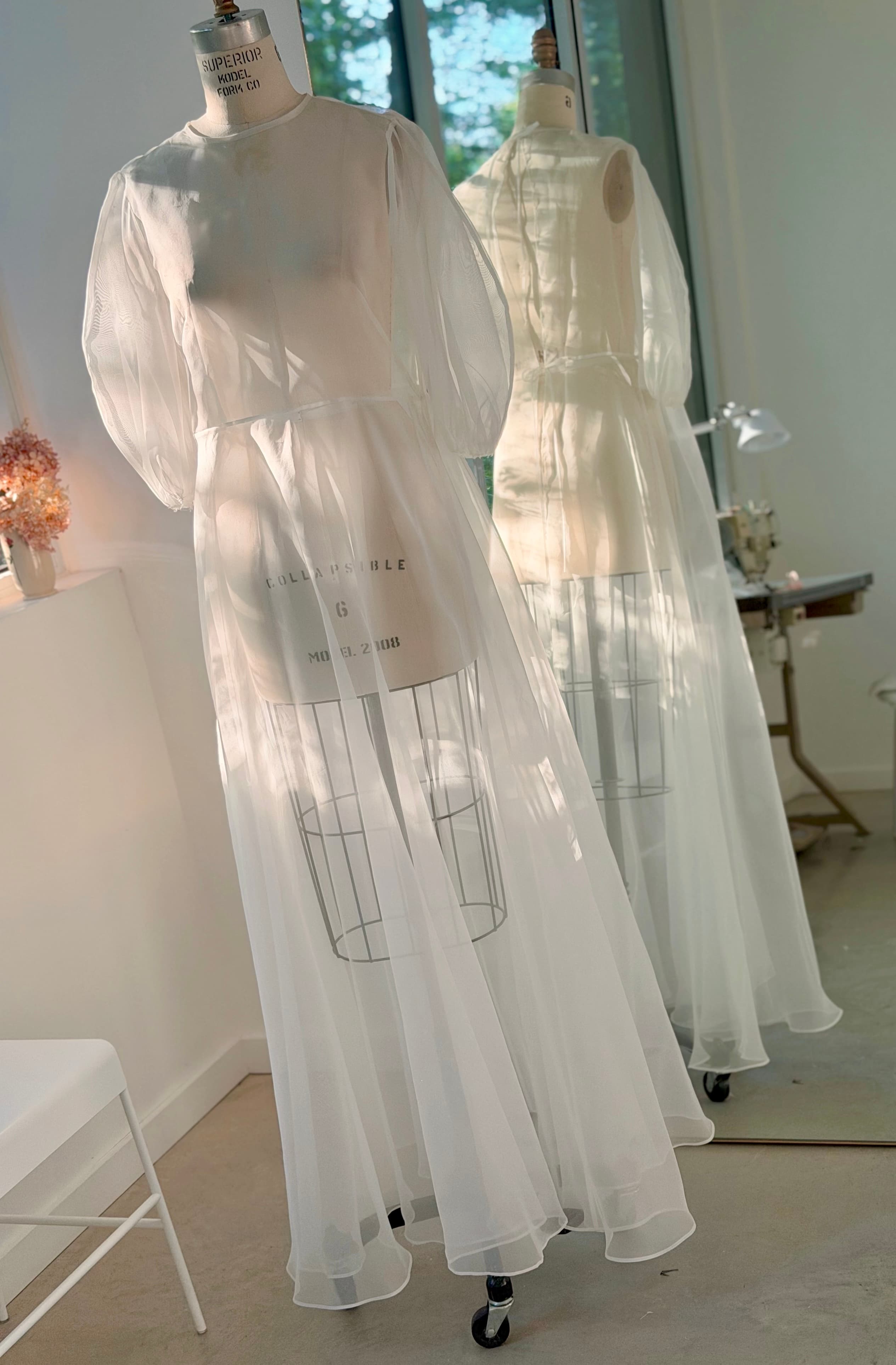 The Maya Dress by Noémiah—a sheer white silk organza dress with long sleeves—is displayed on a mannequin in front of a mirror in a sunlit room, the soft light beautifully highlighting its delicate fabric.