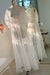 The Maya Dress by Noémiah—a sheer white silk organza dress with long sleeves—is displayed on a mannequin in front of a mirror in a sunlit room, the soft light beautifully highlighting its delicate fabric.