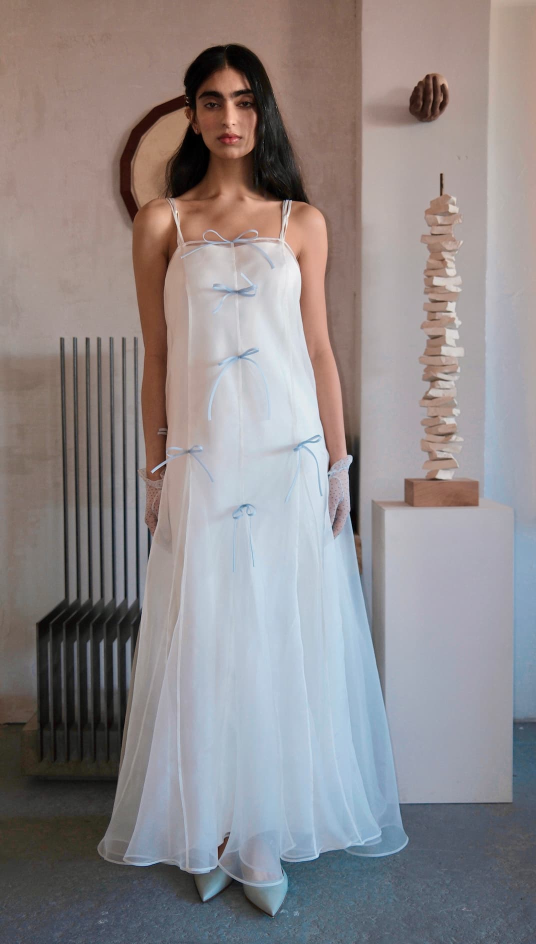 A woman with long dark hair wears the Meg Dress by Noémiah, a sheer organza silk slip adorned with light blue ribbon bows. She stands indoors near abstract art in a neutral, minimalist bridal setting as soft light streams in.