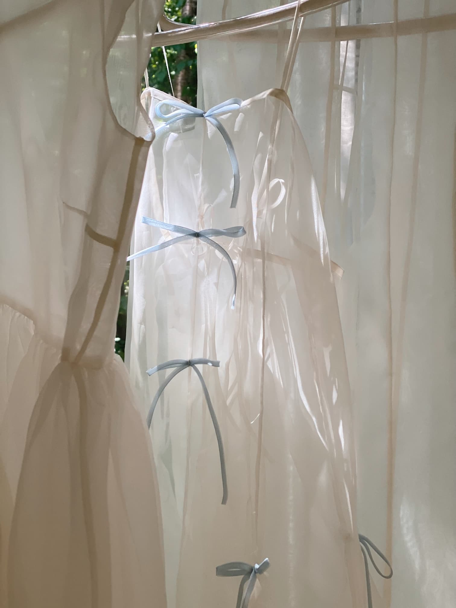 The Meg Dress by Noémiah, a sheer white slip dress in organza silk with delicate blue ribbon ties down the front, hangs on a line with sunlight streaming through. Blurred green leaves form the background.