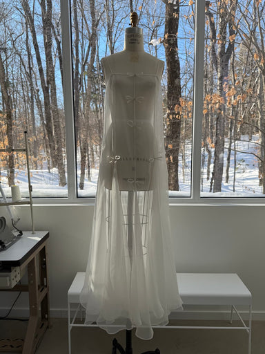The Meg Dress by Noémiah, a sheer white organza silk dress with small bow details, is displayed on a dress form in a bright room with large windows overlooking snowy woods. Sewing machines and benches are nearby.