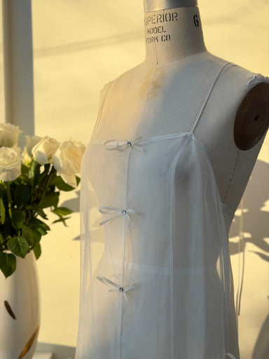 A dress form features the Meg Dress by Noémiah—a sheer, sleeveless organza silk slip with front bows. In the background, a vase of white roses sits as sunlight casts gentle shadows.