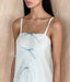 A woman wearing the Meg Dress by Noémiah—a white organza silk slip dress with thin straps and three light blue ribbon bows—stands against a light wooden background.
