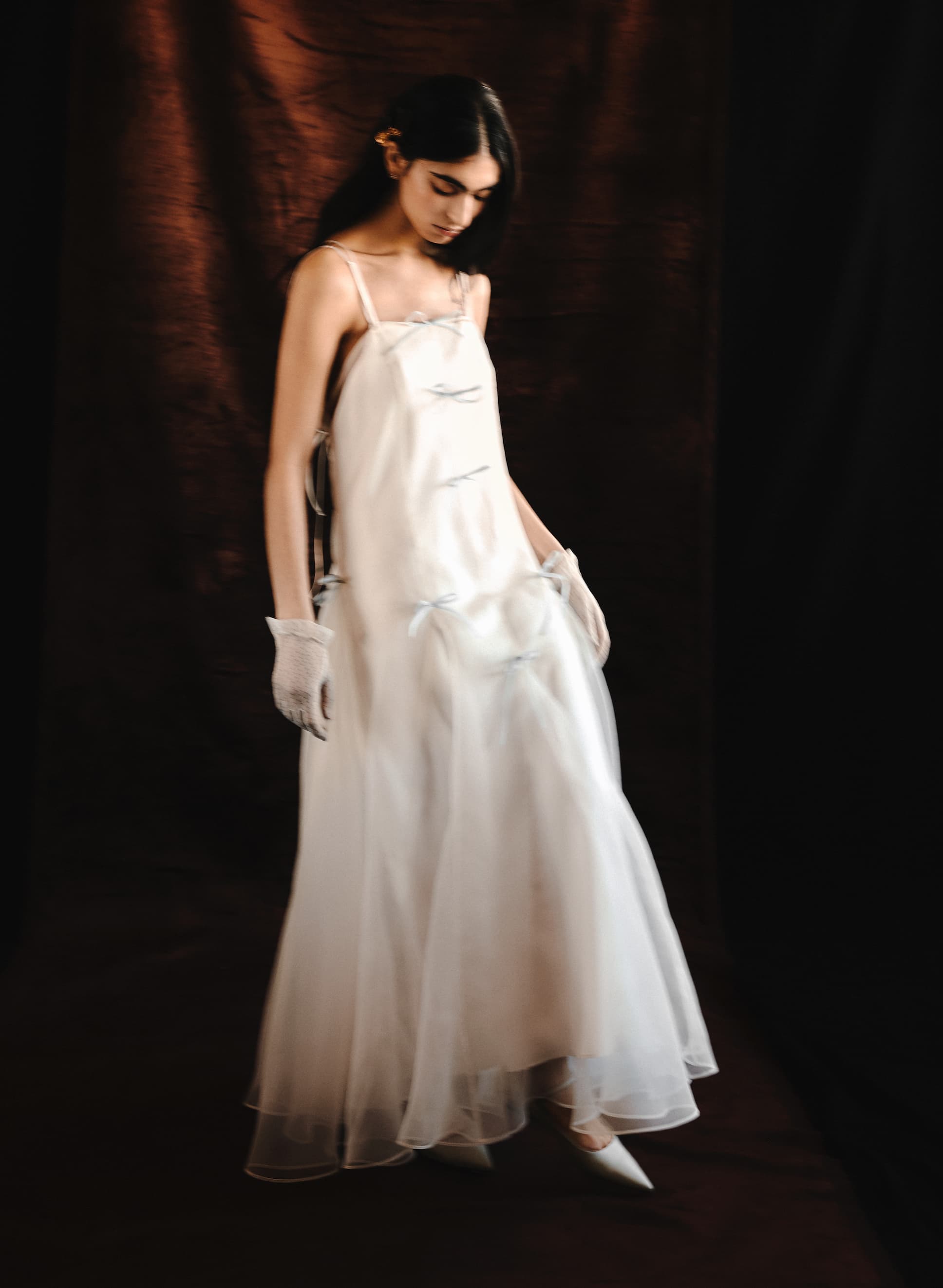 A woman wears the Meg Dress by Noémiah—a long, flowing organza silk slip with thin straps—paired with white gloves. She stands against a dark brown backdrop in a soft, graceful pose. The minimalist bridal image features a subtle blur effect.
