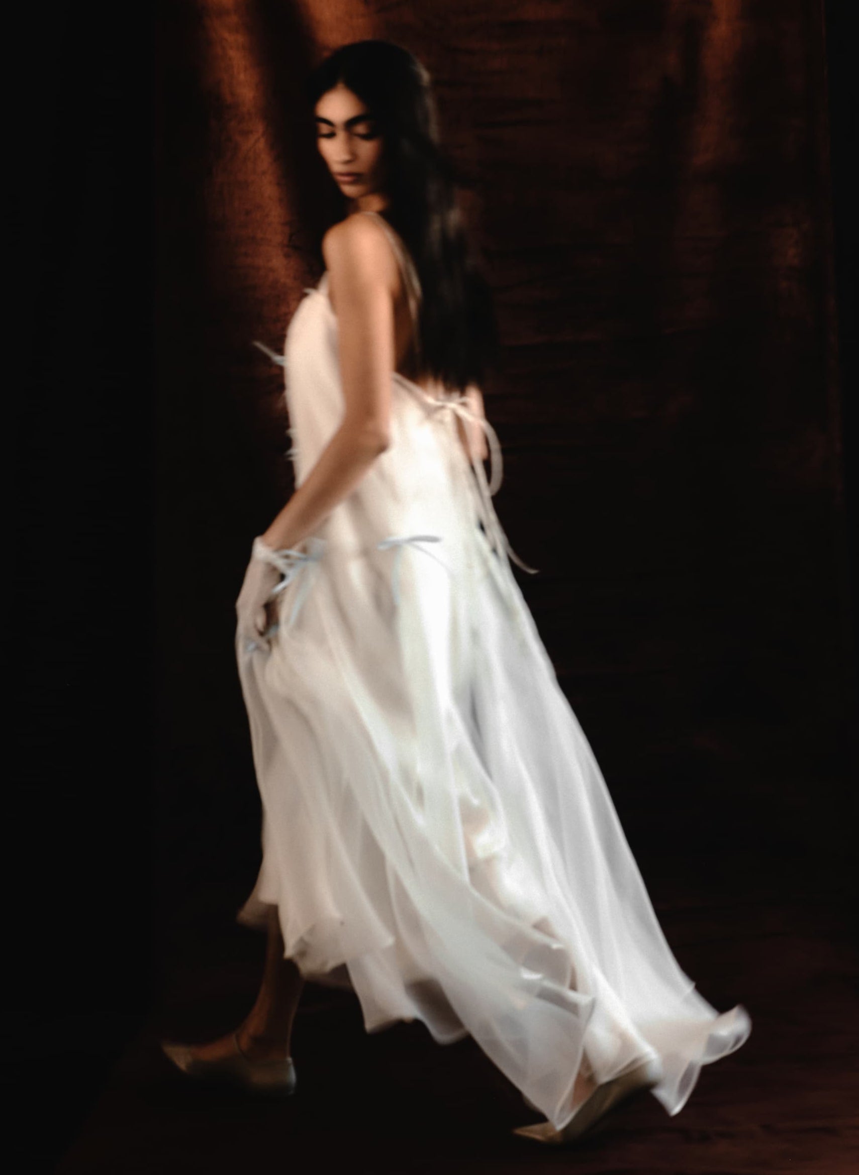 A woman in the Meg Dress by Noémiah, a flowing organza silk slip, walks gracefully against a dark, softly lit background, glancing over her shoulder—an embodiment of minimalist bridal elegance.