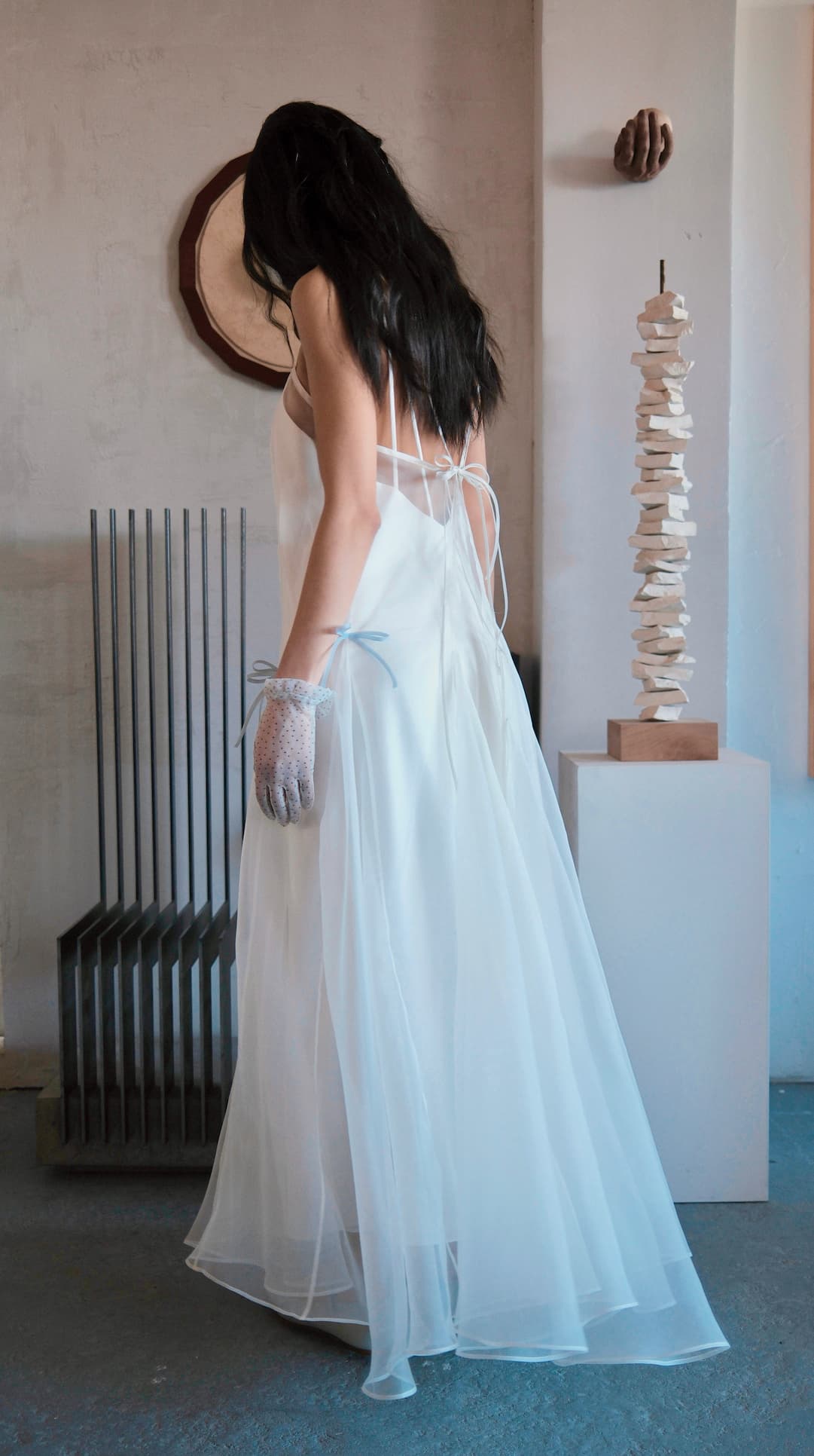A woman with long dark hair stands indoors, facing away, wearing the Meg Dress by Noémiah—a flowing organza silk slip dress with thin straps—paired with sheer gloves. Modern abstract sculptures and a round wall piece complete the minimalist bridal scene.