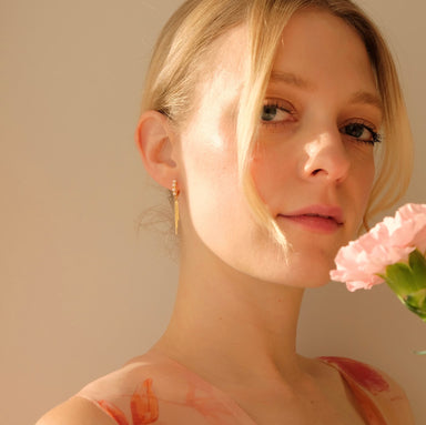 A person with light hair gazes softly at the camera, holding a pink carnation near their face. They wear a floral-patterned top and the Millie Earrings by Noémiah, 14K gold-plated hoops, with gentle, warm lighting highlighting their features.