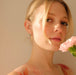 A person with light hair gazes softly at the camera, holding a pink carnation near their face. They wear a floral-patterned top and the Millie Earrings by Noémiah, 14K gold-plated hoops, with gentle, warm lighting highlighting their features.