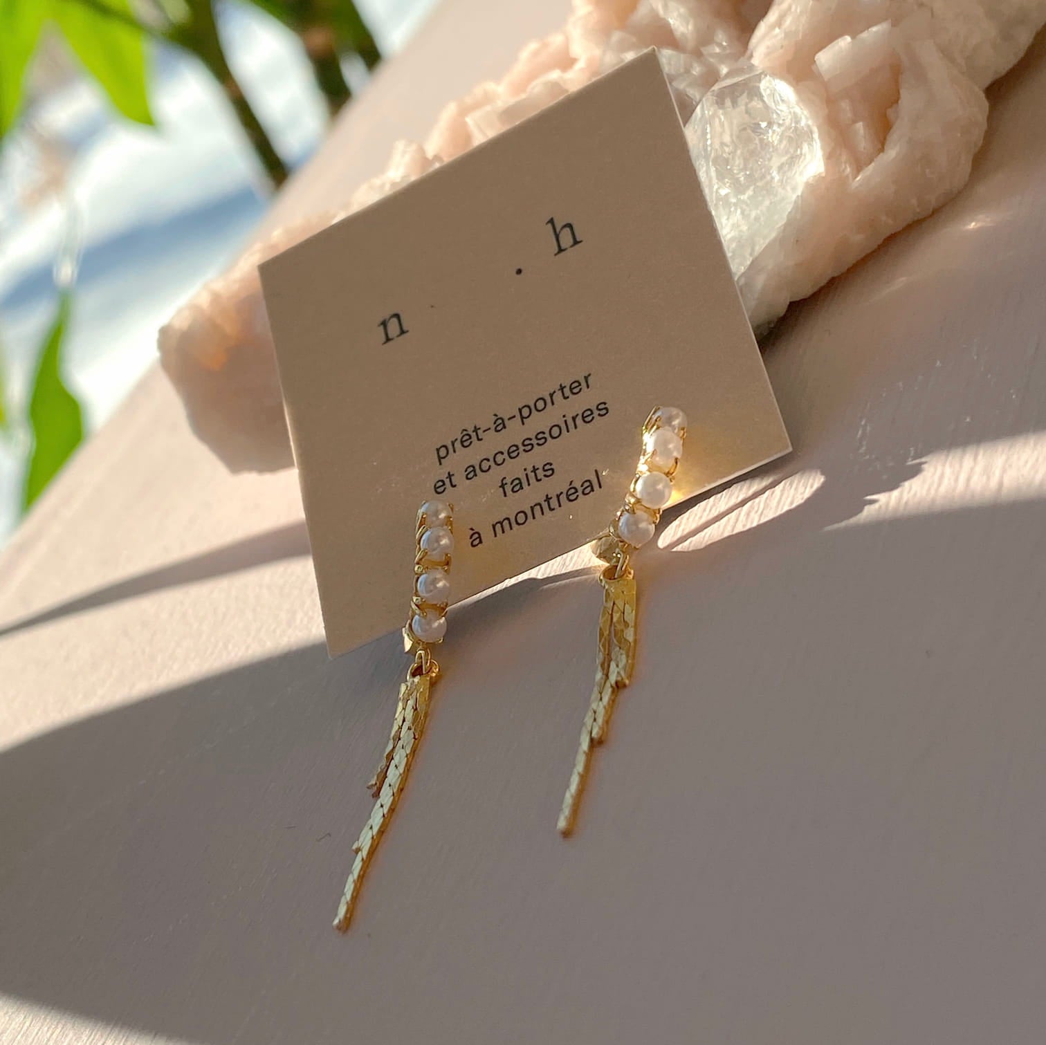 A close-up showcases the Millie Earrings by Noémiah, elegantly displayed on a card. These 14K gold-plated earrings feature small pearls at the top with long, gold dangling elements. The card includes text in French and is set against a textured surface, illuminated by soft natural lighting.