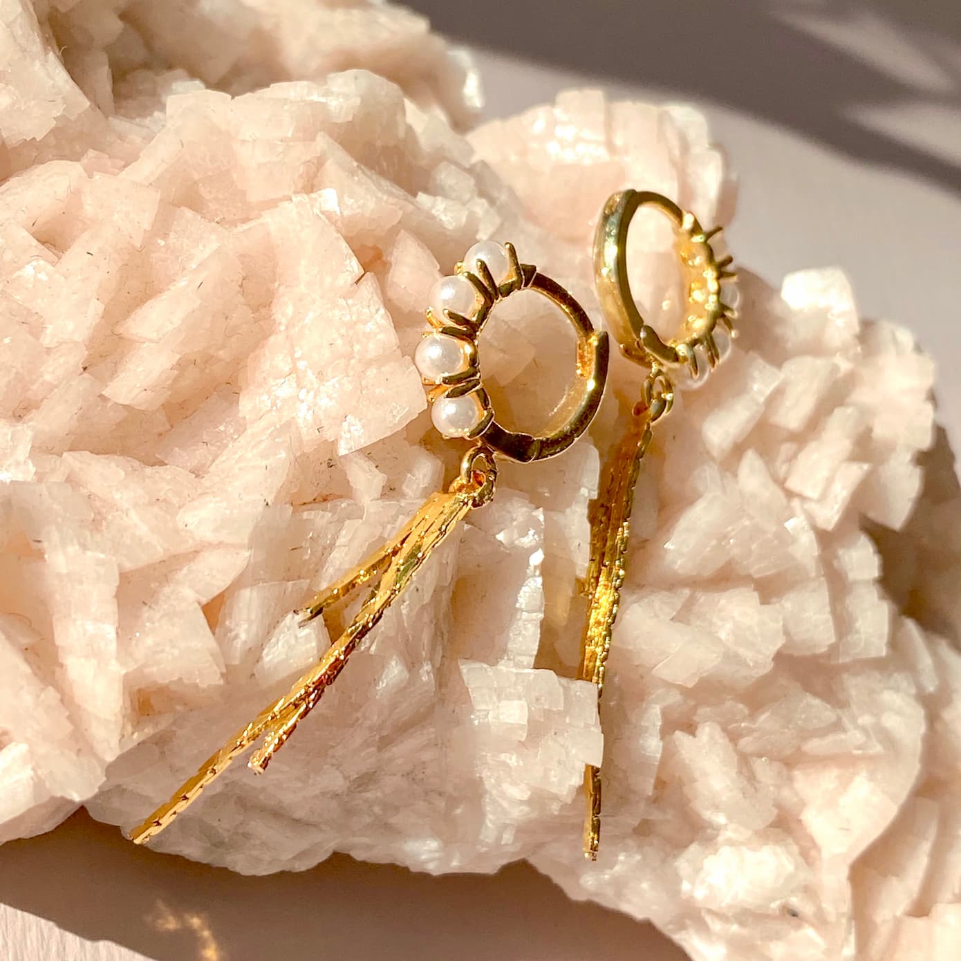 Close-up of the exquisite Millie Earrings by Noémiah, featuring 14K gold plating with pearl accents and textured dangling elements, displayed on a crystalline mineral surface. The earrings catch the light, highlighting their intricate details that evoke classic hoop designs.