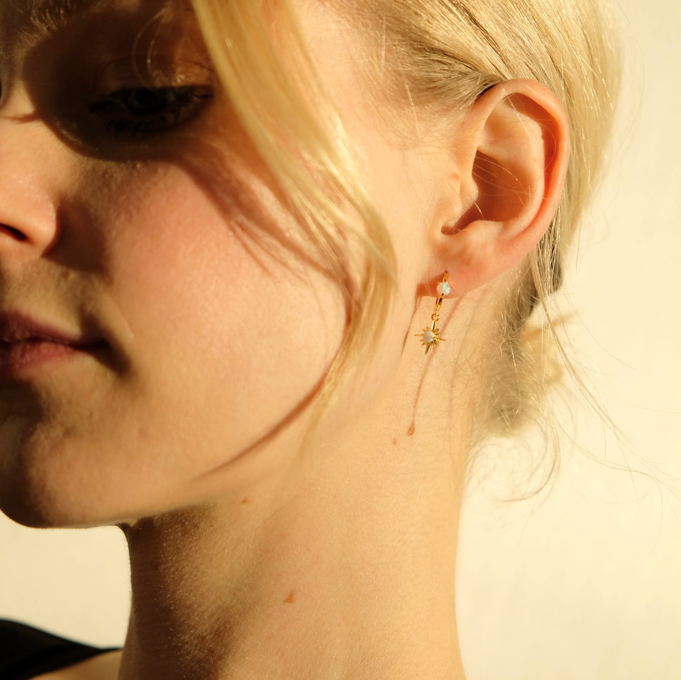 Close-up of a person's face highlighting the Monica Earrings from Noémiah, featuring a golden star shape adorned with a small opal gemstone. Blonde hair partially obscures the face, while 14K gold-plated accents catch the warm lighting, casting soft shadows.