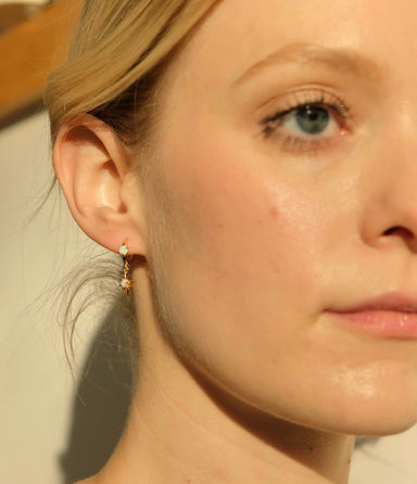 Close-up of a person with fair skin and blue eyes, wearing the Monica Earrings by Noémiah. The softly blurred background highlights their serene expression and the simple elegance of these small, delicate 14K gold-plated hoop earrings.