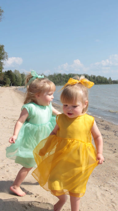 Two young children play on a sandy beach. One wears Noémiahs green dress with the Silk Ribbon — Special Edition in organza, while the other wears a yellow dress with scalloped embroidered edges. The sky is clear with a few clouds, and trees line the background.