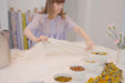 Wearing a light purple dress, an individual carefully arranges sheer fabric on a table adorned with several bowls filled with colorful plant materials, utilizing the Natural Dye Kit by Noémiah to craft natural dyes. Rolled textiles can be seen in the background, while nearby, pink flowers in a vase add charm to this unique scene.