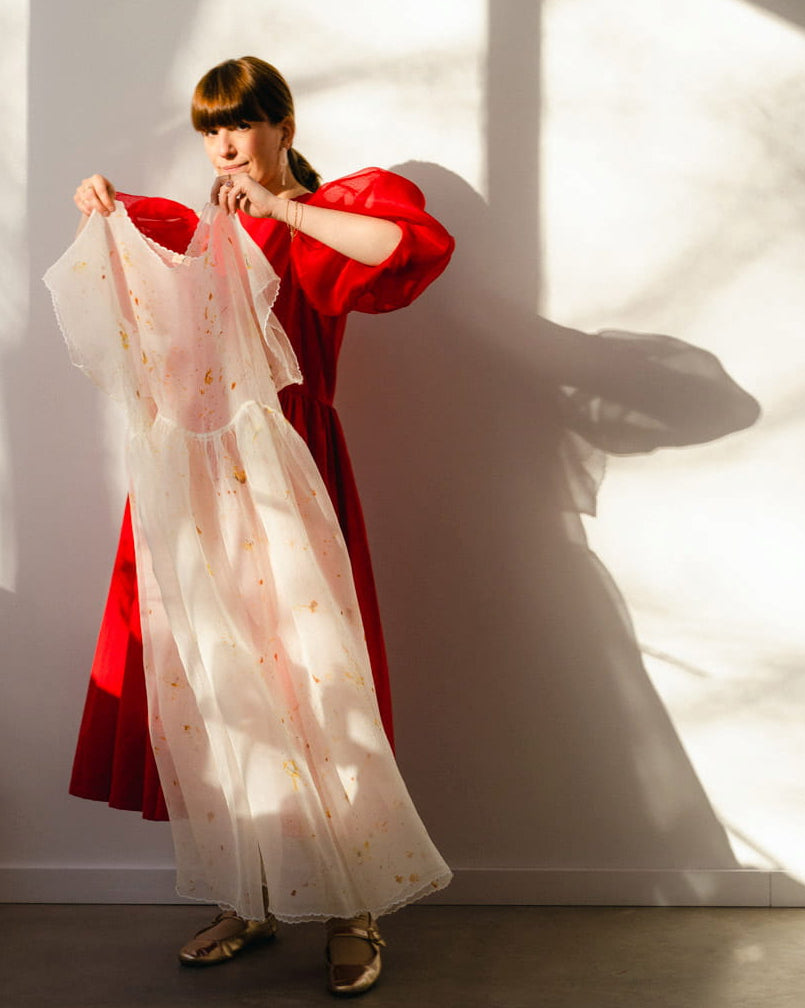 In a sunlit room, a woman wearing gold flat shoes stands holding a sheer white garment adorned with gold accents. She contemplates whether the Noémiah Natural Dye Kit might transform it into something unique. Shadows from the window create patterns on the wall behind her, and a small sculpture sits on a nearby white pedestal.