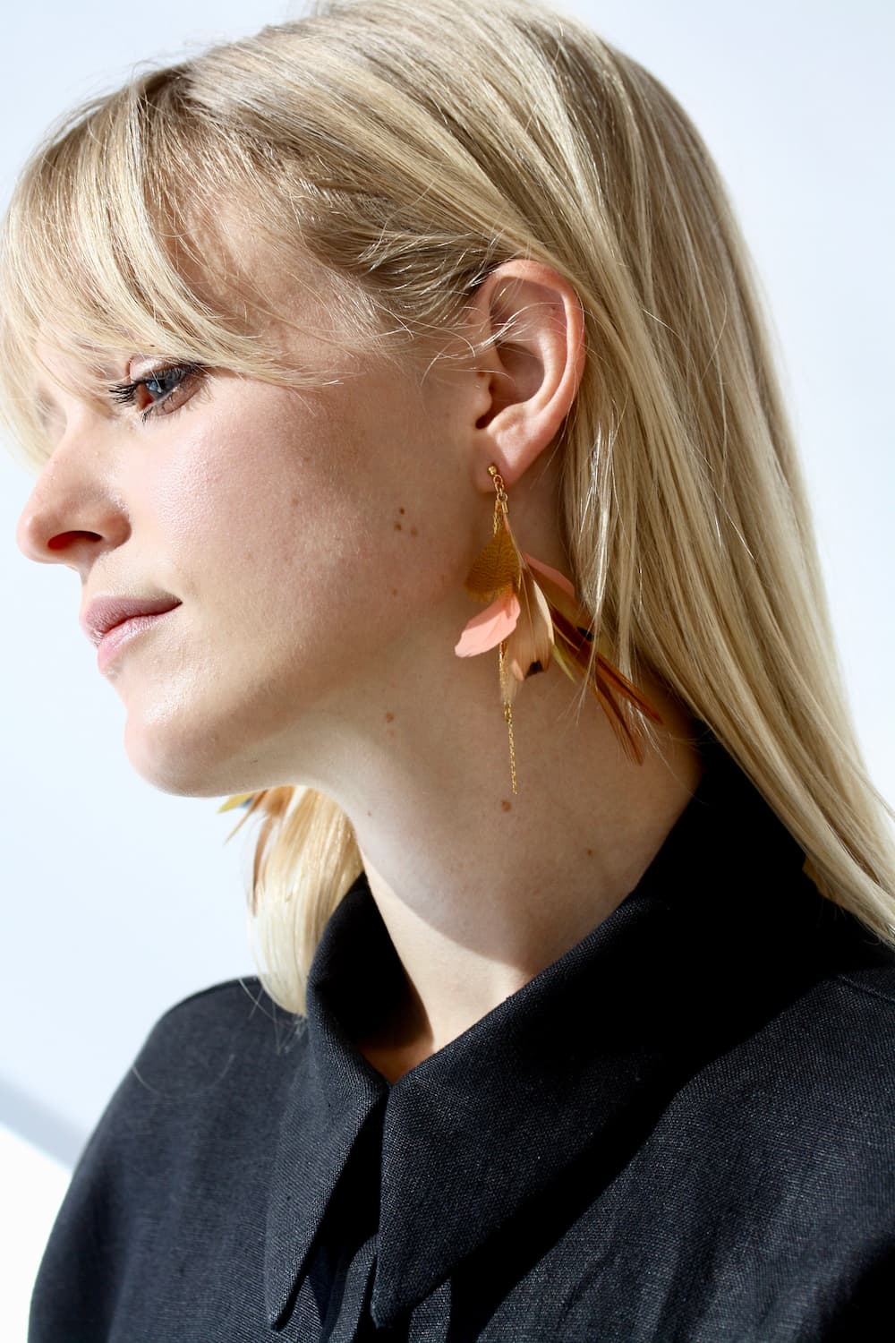 A person with long blonde hair is wearing a black collared shirt and Noémiah's Nicole Earrings, which are feather-shaped clip-ons. They are gazing to the side against a bright background.