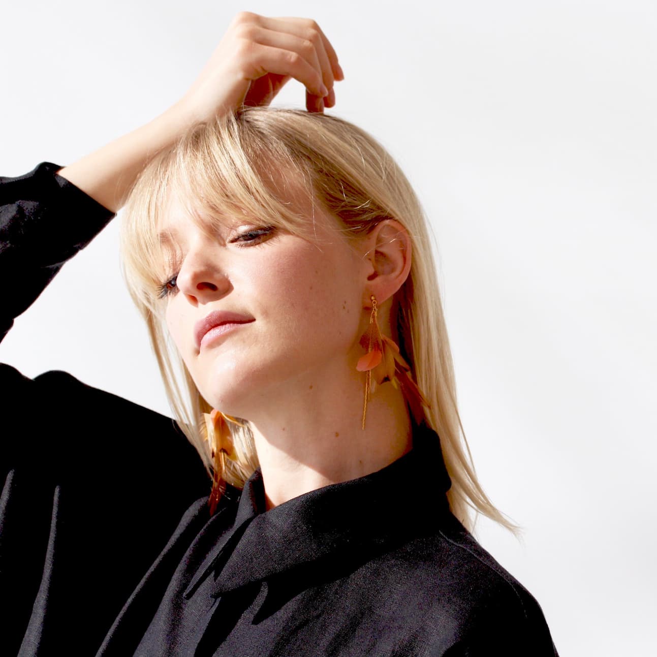 A person with blonde hair wearing large, colorful Nicole Earrings by Noémiah and a black shirt poses with one hand touching their head. The background is white, and the lighting is bright.