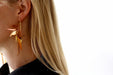 Close-up of a person with long blonde hair wearing the Nicole Earrings by Noémiah, which are clip-on earrings featuring decorative feathers in shades of orange and brown. They wear a black top against a plain white background.