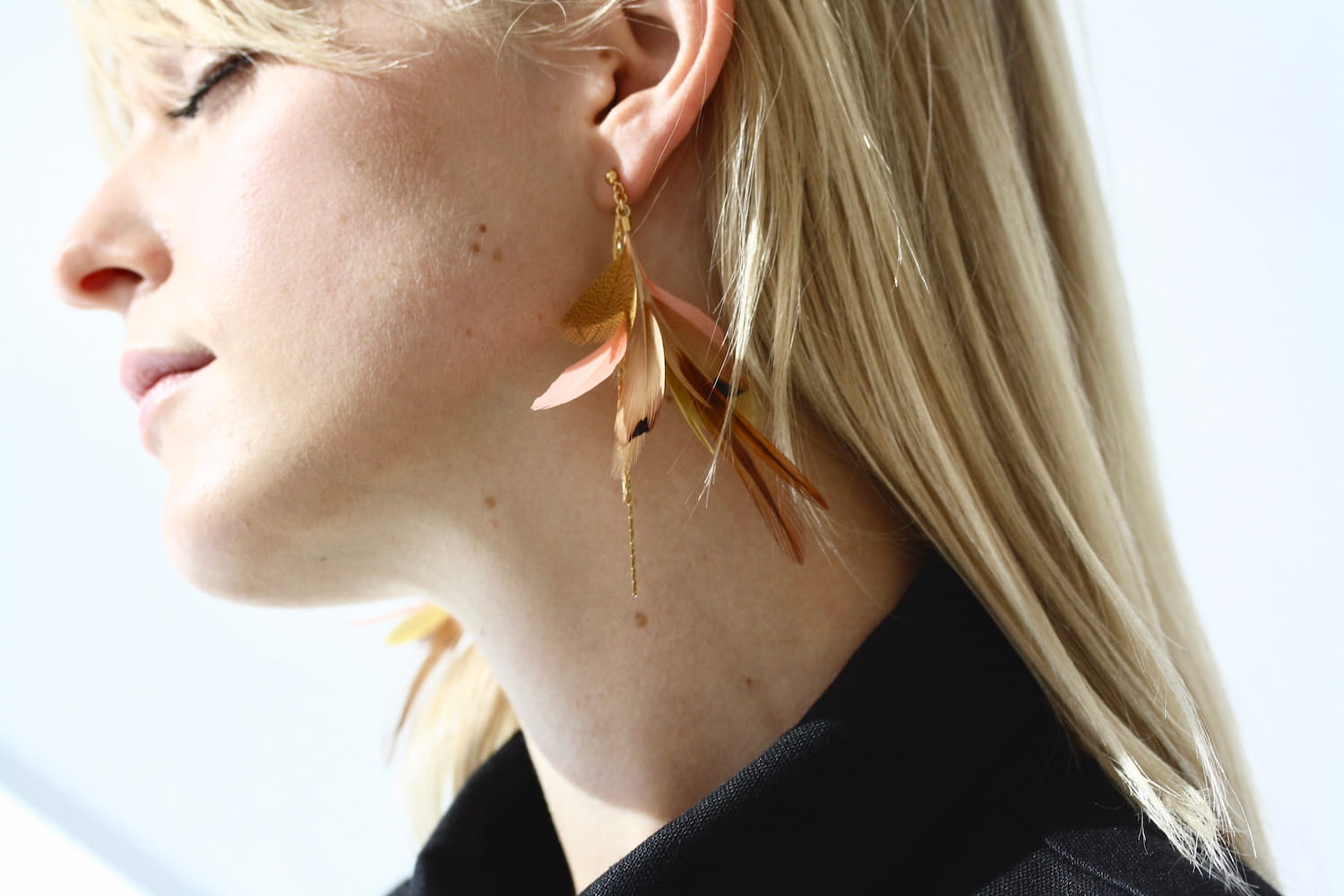 A close-up captures a person with blonde hair wearing the vibrant Nicole Earrings by Noémiah, adorned with gold-filled hooks. The softly blurred background accentuates the earring and the person's smooth skin as they gaze away.