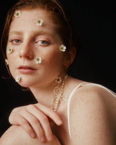 A person with reddish hair and freckles is embellished with small white flowers on their face. They wear the Noémiah Nieve Necklace, a stunning freshwater pearl piece, elegantly draped over a shoulder-baring white top against a dark background.