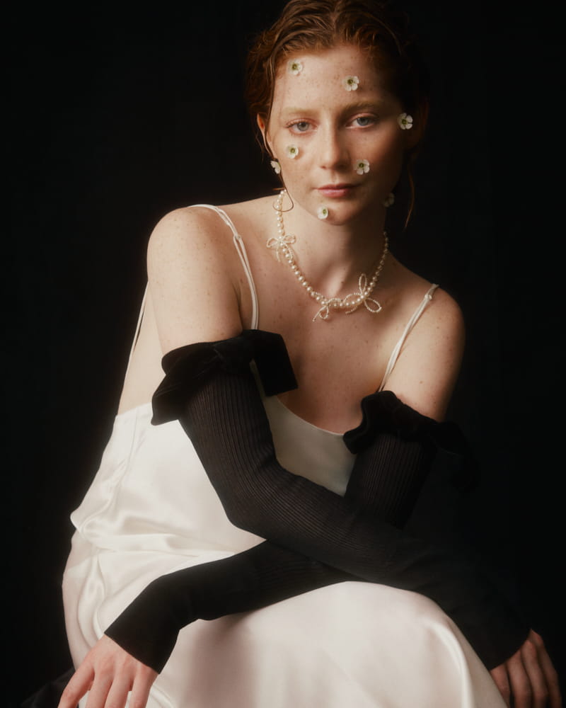 A woman with flowers on her face and the Nieve Necklace by Noémiah, featuring freshwater pearls, around her neck poses against a dark backdrop. She is wearing a white dress embellished with asymmetrical bows and long black gloves, gazing thoughtfully at the camera.