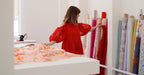 A woman in a red dress browses colorful fabric rolls at Noémiah, holding pink fabric as floral-patterned fabric rests on a nearby table—highlighting the Rush Production Service.